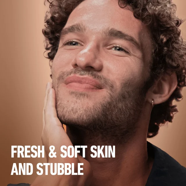 Smiling man with stubble touches face, highlighting fresh & soft skin and stubble.